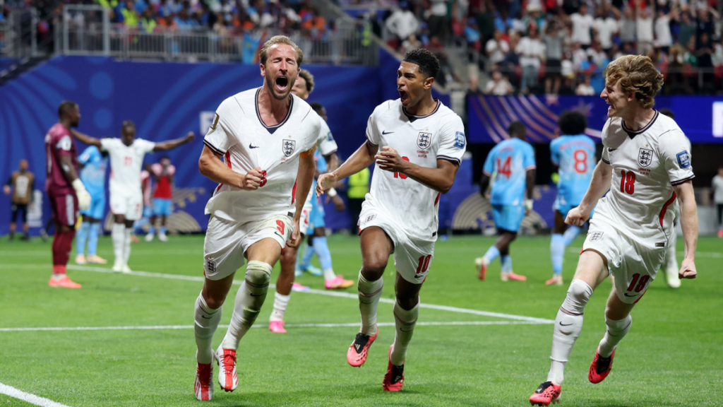 Harry Kane celebra um gol pela seleção da Inglaterra em campo, com o jogador comemorando em primeiro plano e colegas ao redor durante a partida.
