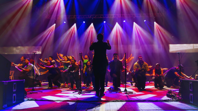Show do Coral Jovem Heliópolis em palco iluminado com luzes coloridas, maestro ou apresentador em destaque de costas à frente de cantores, instrumentos musicais e microfones durante apresentação gratuita em Piracicaba.