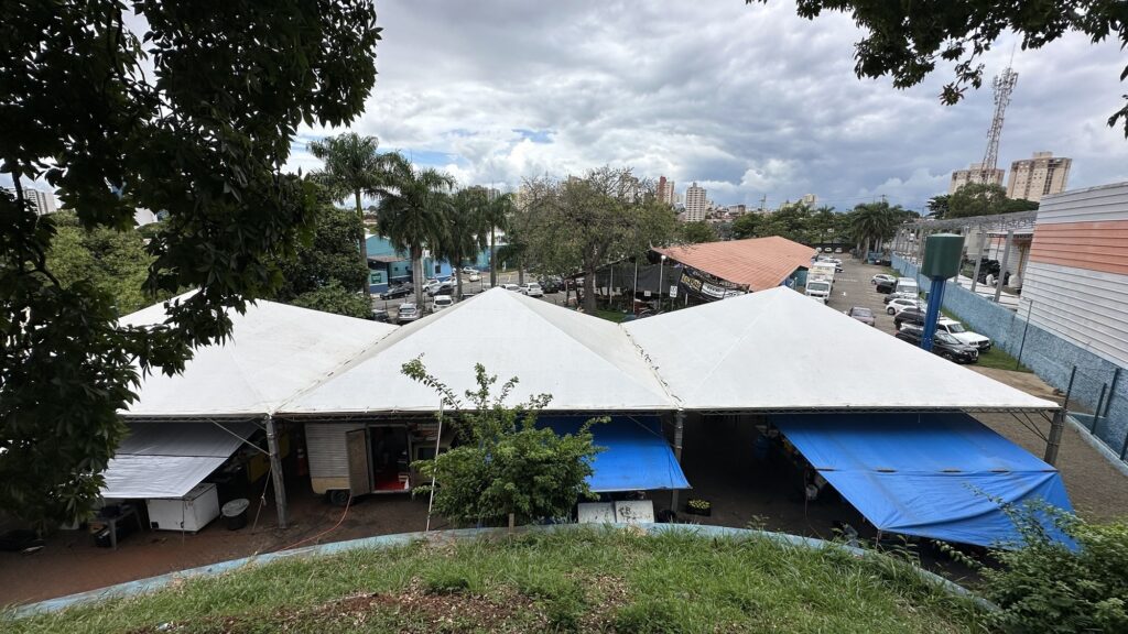 Vista externa de um espaço do Varejão Municipal da Paulista com cobertura metálica e tendas brancas e azuis, com área arborizada e estrutura urbana ao fundo, em Piracicaba.