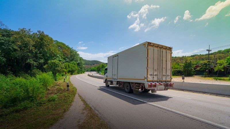Caminhão de carga em rodovia cercada por vegetação, transportando contêiner; cenário que ilustra o frete rodoviário e mudanças aprovadas no Senado.