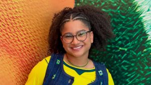 Tia Milena sorri em foto promocional, com cabelo crespo preso para trás e óculos, usando blusa amarela e jardineira jeans, em fundo com luzes laranja e verde