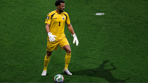 Jogador em campo usando a camisa de goleiro amarela do time, segurando a bola no gramado; destaque para a camisa de Vozinha em leilão mundial que bate recorde.