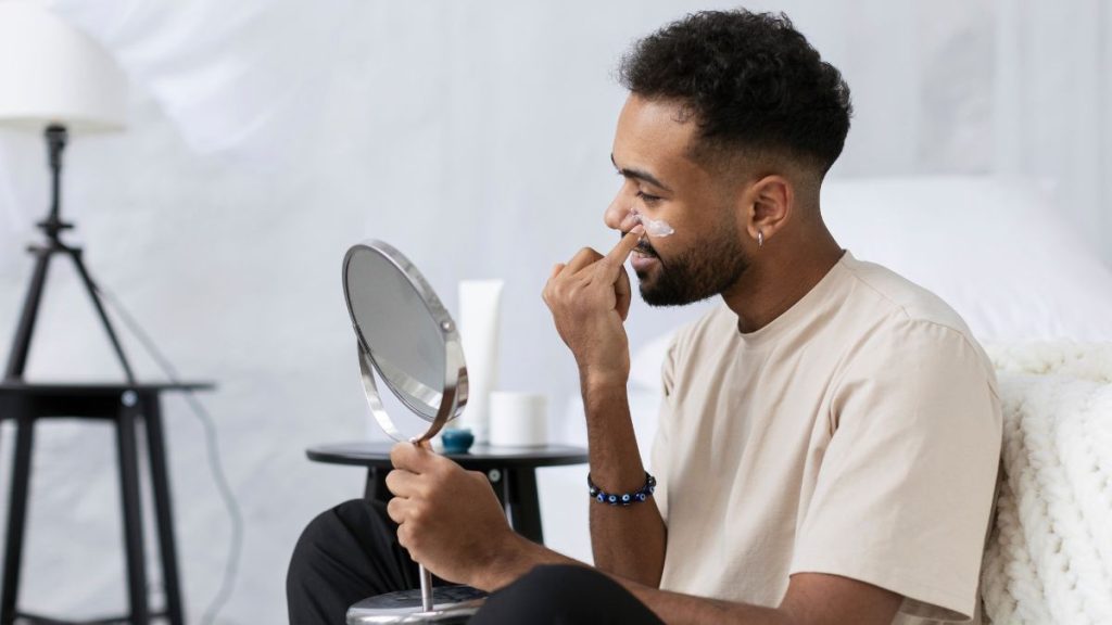 Homem em rotina de skincare masculino em frente ao espelho, aplicando o cuidado no rosto com a mão e fazendo expressão de concentração, em ambiente claro e minimalista.