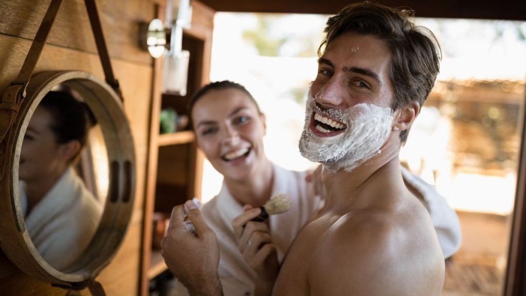 Skincare masculino: homem fazendo a barba com espuma no rosto enquanto se prepara em casa, mostrando uma rotina de cuidados além da lâmina e higiene diária com espelho