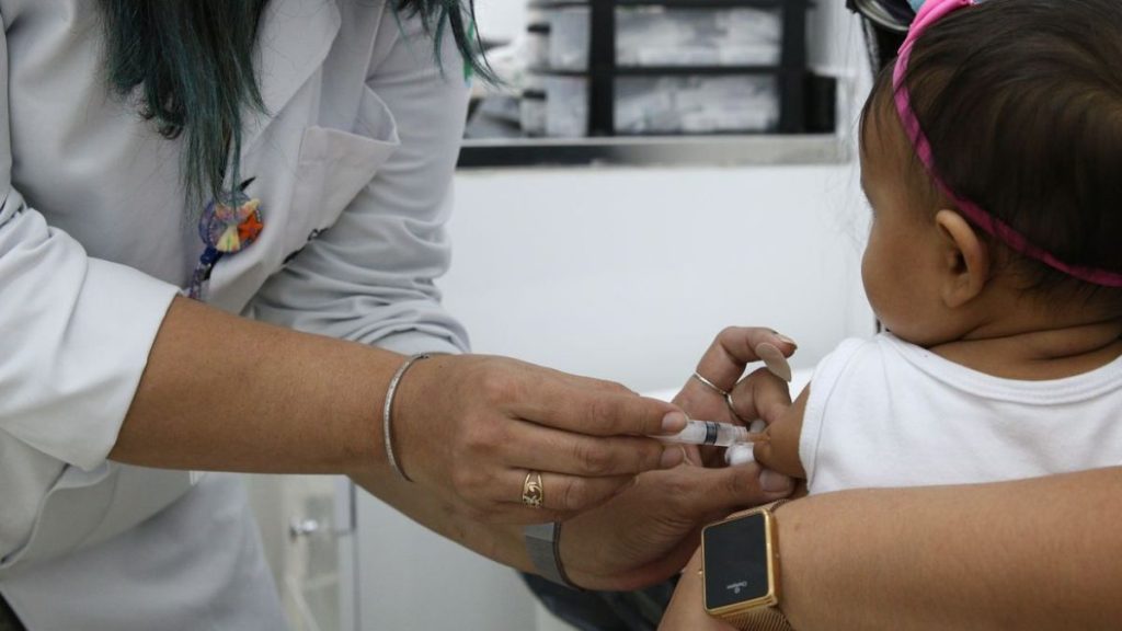 Profissional de saúde aplica uma injeção em uma criança em consultório, destacando crianças sem vacina e a necessidade de ampliar a cobertura no primeiro ano de vida.