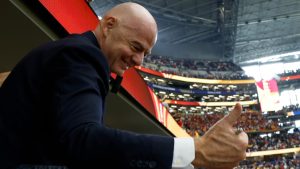 Presidente da Fifa em estádio, em clima de debate, fazendo um gesto com a mão enquanto aparece próximo a uma faixa vermelha e uma torcida ao fundo; imagem usada em notícia sobre denúncia ao COI por suposta violação de neutralidade política.