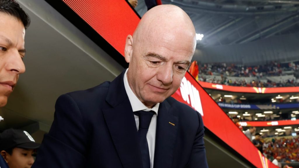 Presidente da Fifa em estádio, sentado e pensativo durante evento esportivo, com bandeira vermelha ao fundo; imagem usada em matéria de denúncia ao COI