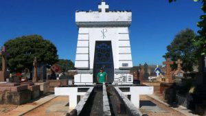 Entrada do Cemitério da Saudade com monumento branco ao centro, jardins e lápides ao redor sob céu azul, durante visita guiada no Cemitério da Saudade.