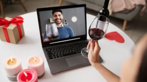 Casal brindando com taças de vinho em uma mesa enquanto conversa por vídeo em um notebook, sugerindo juras de amor e união estável virtual a partir de um aplicativo