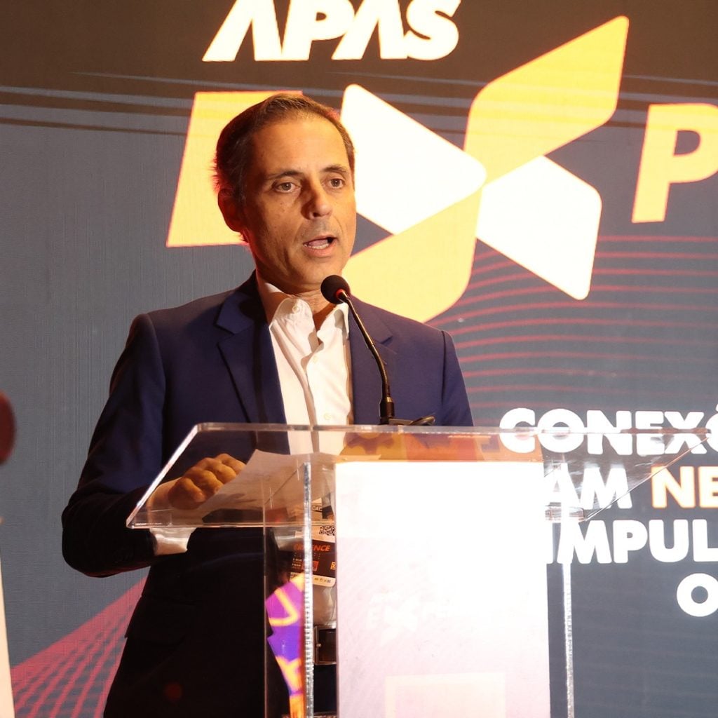 Erlon Ortega, presidente da APAS, discursando em um palco na APAS Experience 2026, ao lado de um púlpito de acrílico com microfone e com fundo do evento.