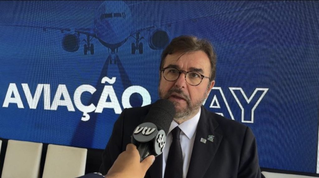 Diretor-presidente do IBI, Mário Povia, durante entrevista em evento de aviação day, ao lado de painel com a frase “AVIAÇÃO DAY” e em frente a microfones