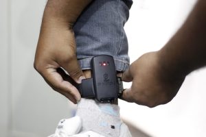 Técnico ajustando uma tornozeleira eletrônica para monitoramento de agressor, com o dispositivo preso ao tornozelo. Projeto de comissão e aprovação para obrigar pagamentos.