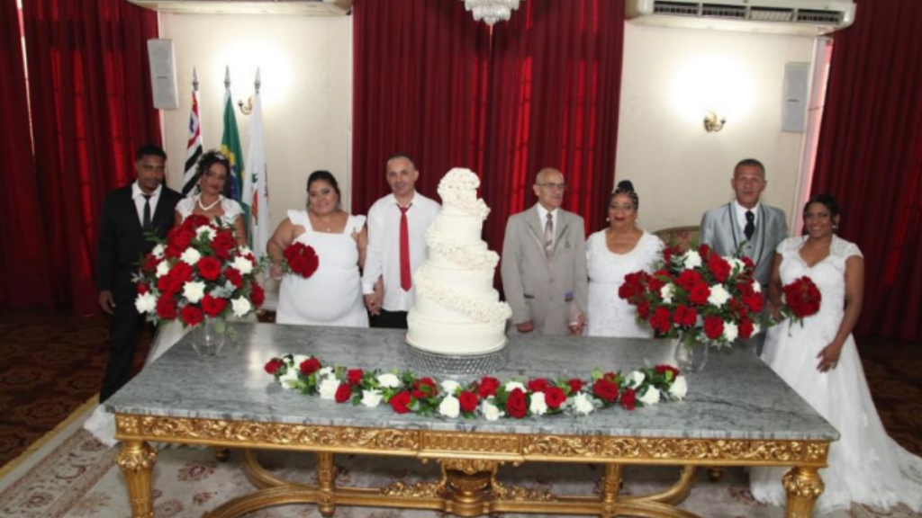 Casamento Comunitário em Santos: moradores posam em cerimônia com mesa decorada e bolo de noiva ao centro, usando trajes formais e buquês de rosas vermelhas, em sala com cortinas e decoração oficial. Crédito: Carlos Nogueira / Prefeitura de Santos.