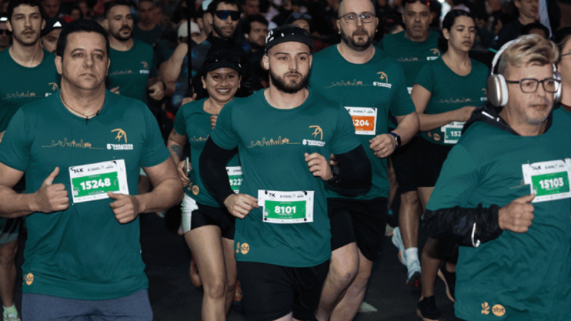 Corredores usando camisetas verdes seguram os kits com números de participação durante a retirada de kits da Maratona de Campinas, com participantes correndo em pista sob iluminação noturna.