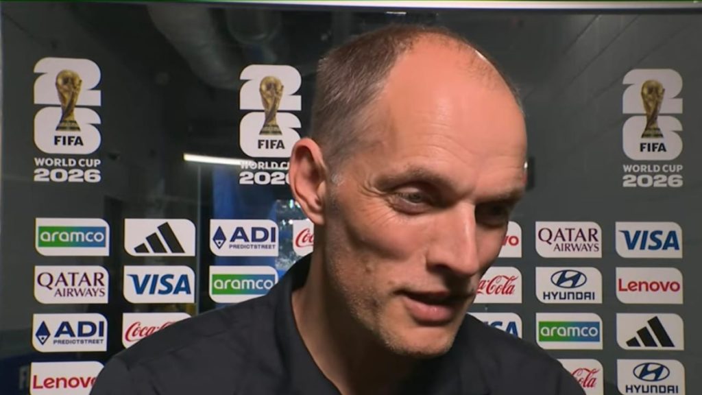 Thomas Tuchel em coletiva de imprensa, em frente ao painel de patrocinadores da FIFA World Cup 2026, com expressao concentrada e iluminação de entrevista