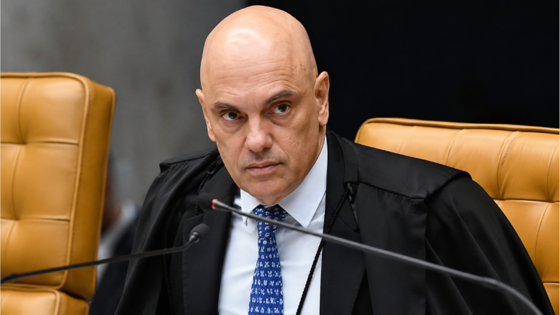 Alexandre de Moraes no plenário do STF, em foto de arquivo, sentado em cadeira de couro e usando toga preta com faixa azul durante sessão, com microfones ao fundo.