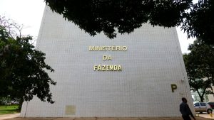 Prédio do Ministério da Fazenda ao ar livre, com árvores ao redor, destacando a fachada que informa “MINISTÉRIO DA FAZENDA”. Contexto ligado à inflação 2026, com governo elevando projeção para 5,1% e prevendo estouro da meta.