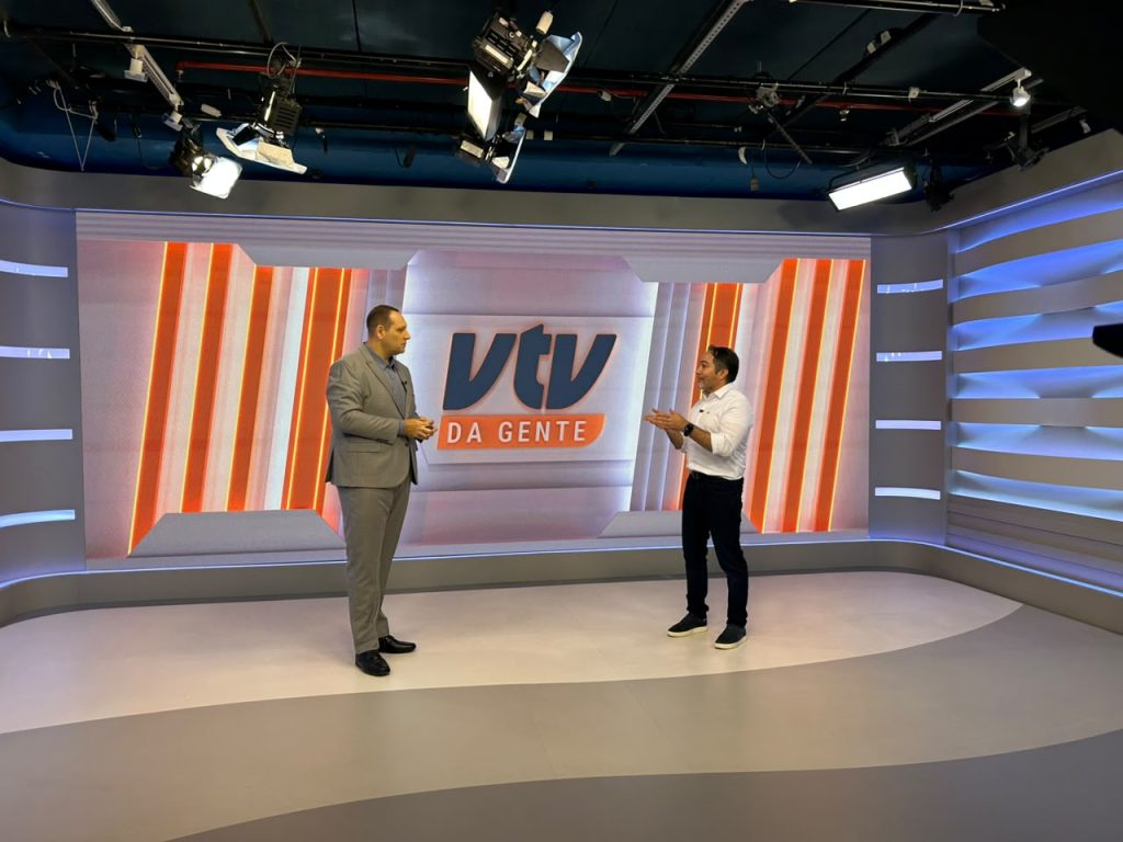 Carlos Piani visita a sede da VTV SBT em estúdio de TV com iluminação profissional e telão “VTV da Gente”, em foto durante encontro na Baixada Santista.