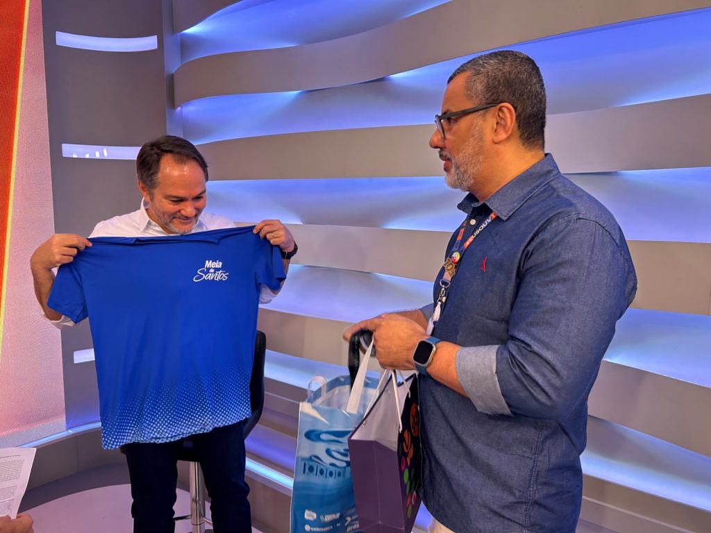 Carlos Piani durante visita à sede da VTV SBT, ao lado de colaboradores, entregando uma camisa da campanha “Meia Santos” em evento na Baixada Santista.