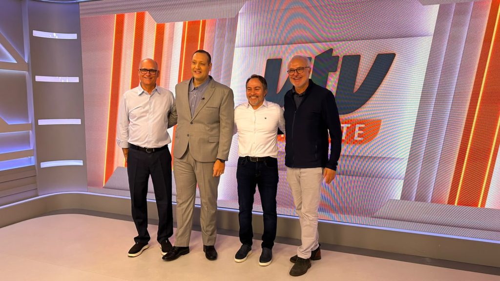 Carlos Piani aparece posando ao lado de quatro homens na sede da VTV SBT, em cenário de estúdio com fundo laranja e branco. Foto de visita institucional.
