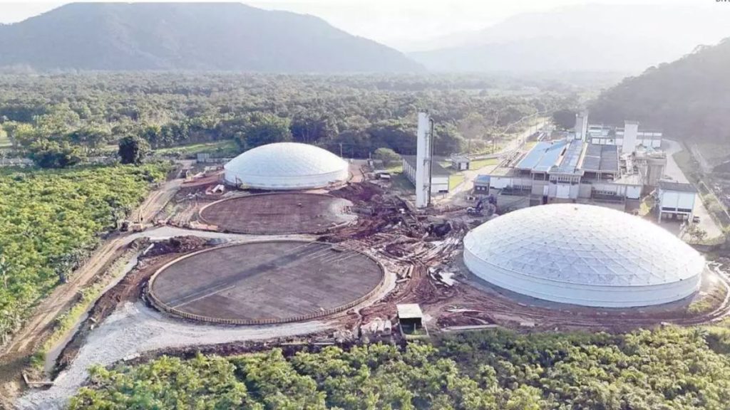 Vista aérea de obras da Sabesp Baixada Santista com novas estruturas de reservação e tanques de armazenamento em área industrial ao lado de vegetação e montanhas.