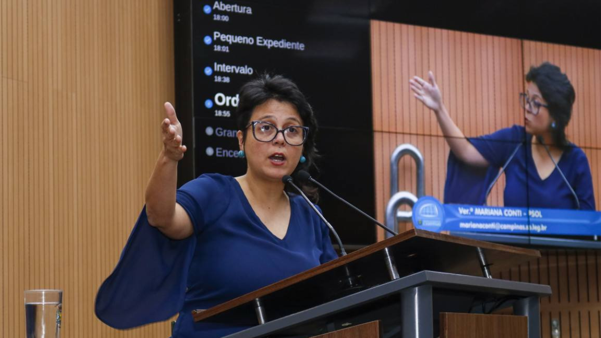 Sessão em plenário com Mariana Conti ao microfone, em audiência, levantando a mão para se manifestar; ao fundo, outra pessoa em painel, tema do caso Vini Oliveira.