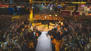 Vista de um grande palco durante a CCXP26, com multidão lotando o evento de cultura pop e banners de grandes marcas como Warner Bros e Barbie, ideal para divulgação de ingressos CCXP26.