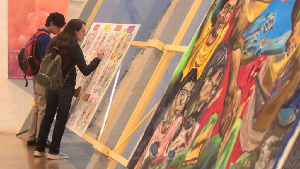 Visitantes apreciam exposição de artes indígenas em museus de Campinas, com painéis e pinturas coloridas sobre rituais do Brasil, durante o evento “Bancos Indígenas do Brasil – Rituais”.