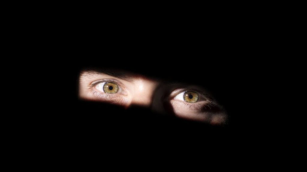 Imagem em close mostra olhos observando através de uma abertura, com fundo escuro. Visual sugere o comportamento de um suspeito de filmar mulheres em banheiro em Santos, sem consentimento.