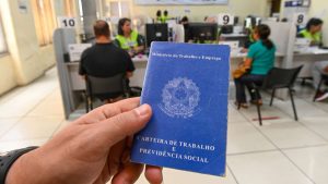 Mão segurando a Carteira de Trabalho e Previdência Social em ambiente de atendimento, com pessoas ao fundo; imagem representa vagas de emprego em Campinas com salários de até R$ 5,1 mil pelo CPAT Campinas.