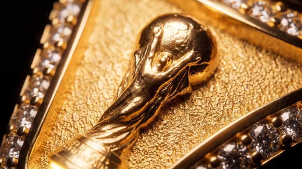 Close-up do anel da FIFA com a reprodução da taça da Copa do Mundo em acabamento dourado, destacando o detalhe do metal em fundo texturizado e brilho característico.