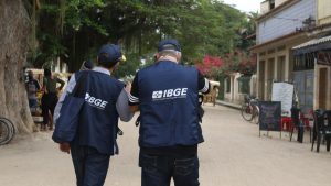 Concurso do IBGE: agentes do IBGE em uniforme azul, vistos de costas, em frente a prédio durante trabalho de campo
