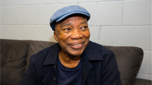 Milton Nascimento em foto de entrevista sentado em um sofá, usando boina azul e jaqueta escura, em ambiente com parede clara ao fundo.