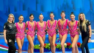 Equipe da seleção brasileira de ginástica rítmica em uniforme rosa, posando sorridente em pista de competição ao lado de comissão técnica, em foto de convocação para o Mundial.