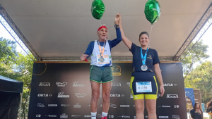 Pódio da Maratona de Campinas com dois atletas comemorando a vitória sob a tenda do evento, com medalhas no pescoço e números de corrida expostos. Muito além da corrida, conheça histórias inspiradoras da Maratona de Campinas.
