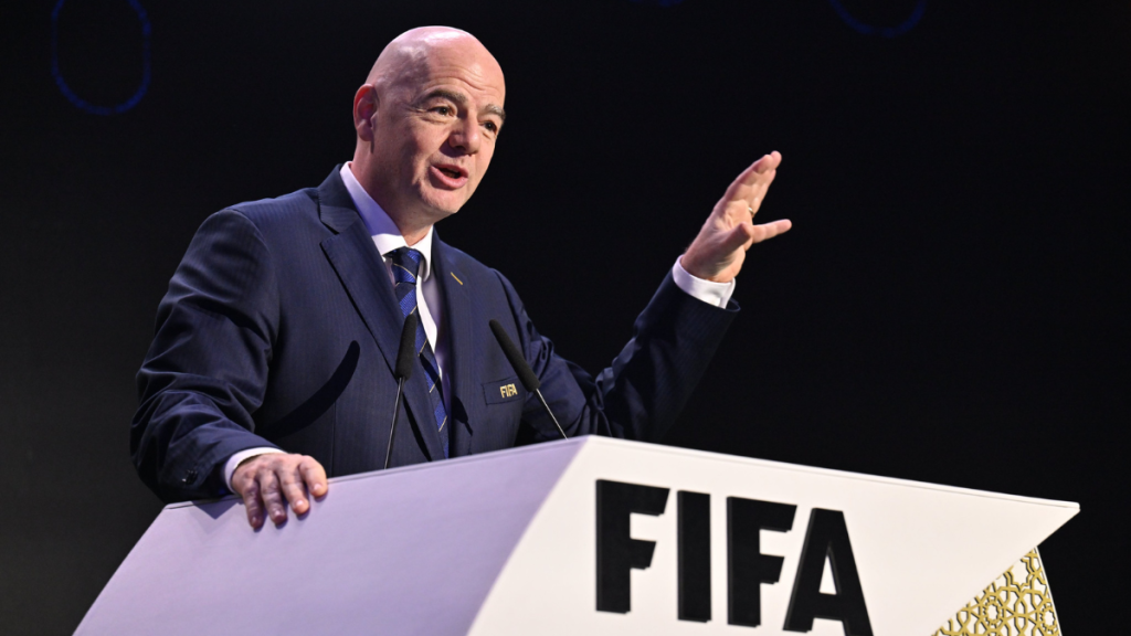 Gianni Infantino, presidente da Fifa, discursando em um evento, com as mãos erguidas atrás de um púlpito com o logo da FIFA ao fundo. Foto de Reprodução.