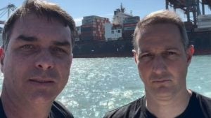 Flávio Bolsonaro ao lado de outra pessoa em visita ao Porto de Santos, com navios e contêineres ao fundo em cenário de área portuária.