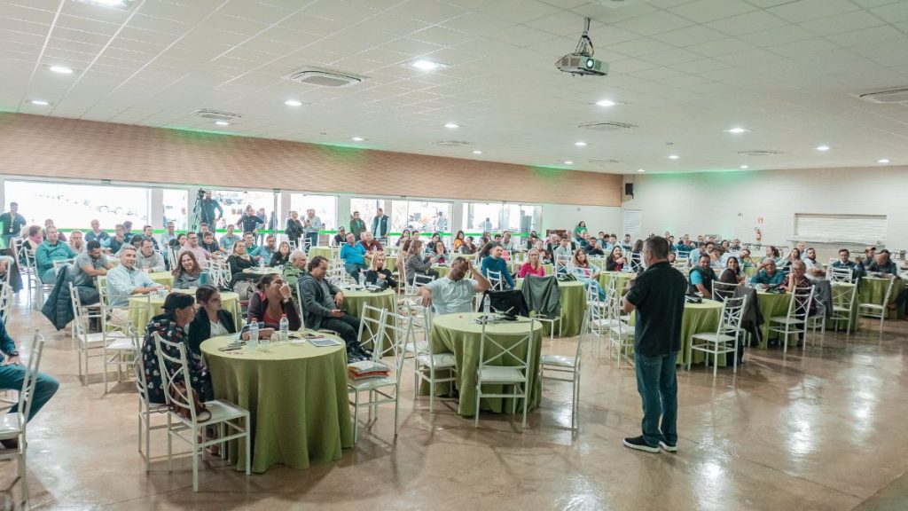 Salão de evento com público sentado em mesas e palestrante em pé, em encontro gratuito nesta quarta-feira com palestras sobre clima, finanças e mercado de grãos, coplacana