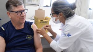 Campinas libera vacina pneumocócica: profissional de saúde aplica injeção no braço de uma pessoa durante atendimento em sala de vacinação, com caixa de insumos ao fundo.