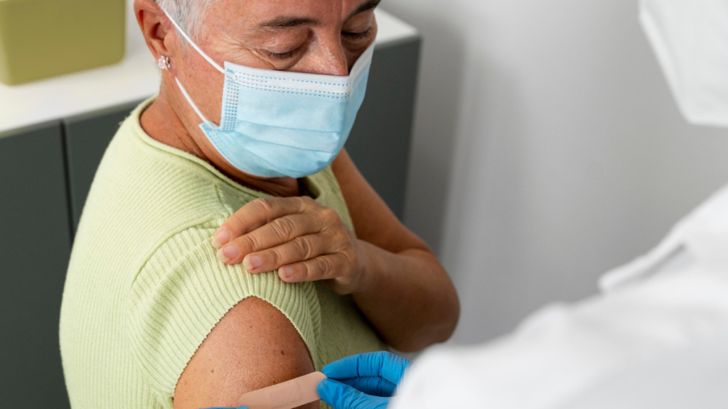 Vacina pneumocócica: Profissional de saúde aplica uma vacina em uma idosa usando máscara facial e luvas azuis, em ambiente clínico; ampliação temporária da vacinação para aumentar a cobertura e evitar desperdício de doses.