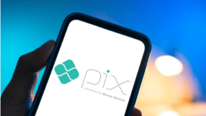 Pix alvo dos EUA: mão segurando smartphone com ícone do sistema de pagamentos instantâneos Pix na tela, com fundo azul desfocado.