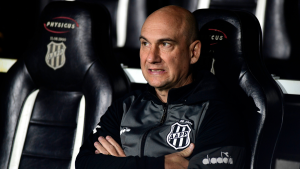 Zanardi Ponte Preta posa no banco de técnico, sentado ao lado dos escudos do clube em um ambiente de comissão técnica, com expressão séria durante partida.