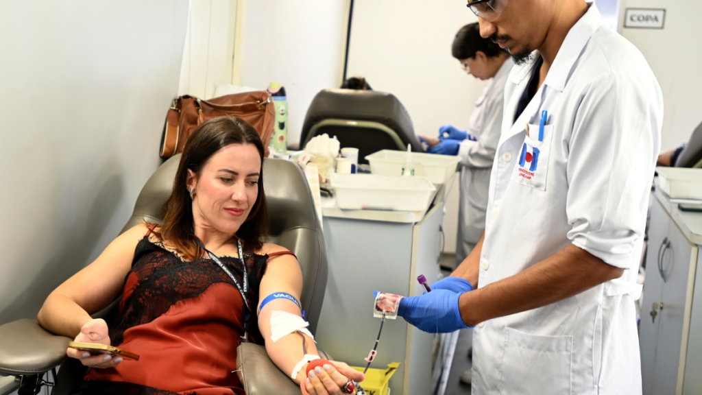 Coleta de sangue em Campinas em unidade móvel: mulher em cadeira de doação enquanto profissional de saúde prepara amostra com material esterilizado. Ação para aumentar doações na região.