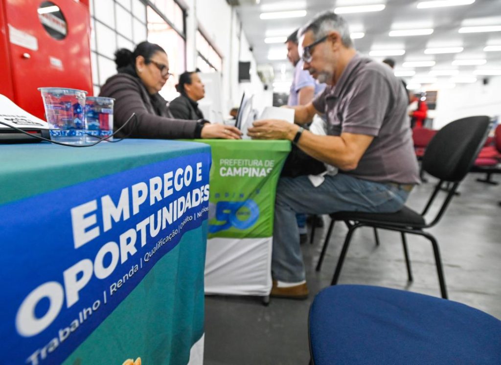 vagas de emprego campinas: Feirão de emprego em Campinas: participantes conversam com recrutadores em sala de evento, com mesa azul e verde escrito “EMPREGO E OPORTUNIDADES” e banca de atendimento.