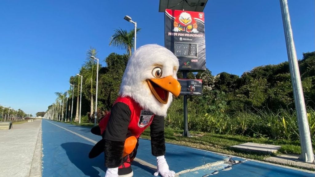 Parque de Lazer com radar de velocidade: mascote de pavão na calçada ao lado de um radar exibindo velocidade em um dia ensolarado.