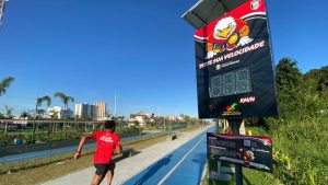 Parque de Lazer com radar de velocidade: Imagem mostra um corredor em pista com sinalização ao lado de um equipamento com painel de “teste sua velocidade” e indicação de velocidade em km/h, em parque ao ar livre.