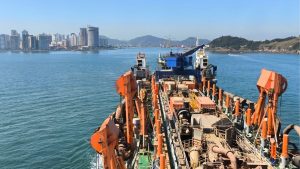 Aproximação de uma embarcação de dragagem no mar durante o aprofundamento do canal do Porto de Santos, com porto e arranha-céus ao fundo.