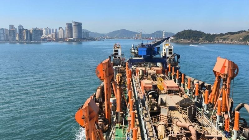 Aproximação de uma embarcação de dragagem no mar durante o aprofundamento do canal do Porto de Santos, com porto e arranha-céus ao fundo.