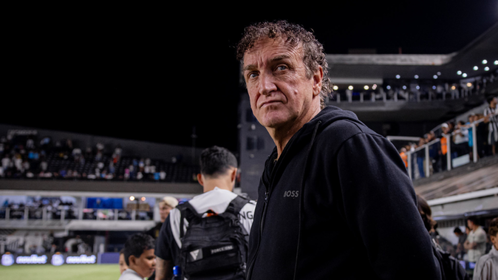 Cuca, técnico do Santos FC, aparece em foto no estádio durante partida, usando moletom escuro e expressão concentrada, com arquibancadas ao fundo.