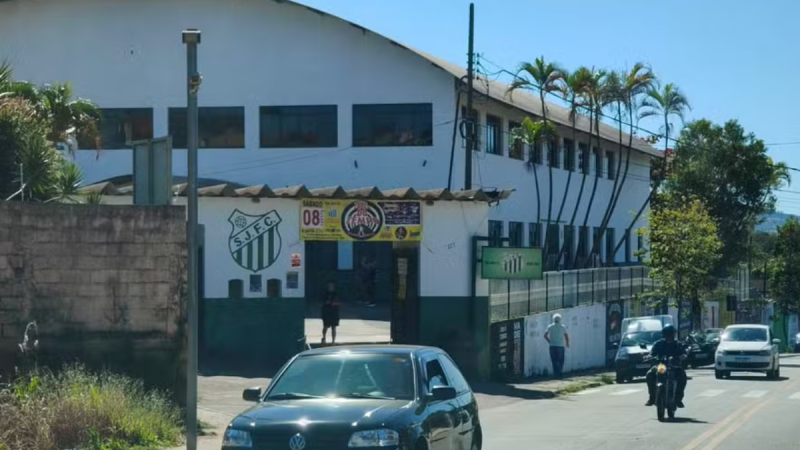 racismo e homofobia nos Jogos Regionais: Vista externa de um complexo esportivo no qual um atleta Guarani denuncia racismo e homofobia, registra BO e é eliminado dos Jogos Regionais. Ao fundo, veículos e um prédio de estádio.